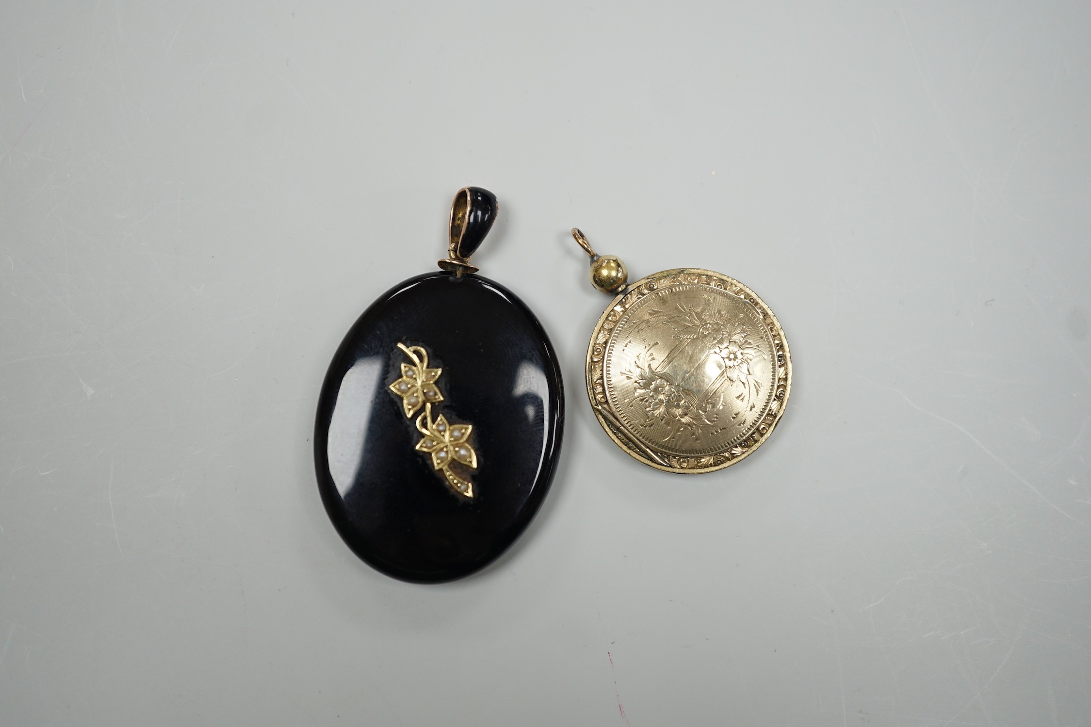 A Victorian yellow metal and seed pearl mounted black onyx oval pendant locket, with enamelled bale, overall 53mm and an engraved gold plated circular pendant locket.